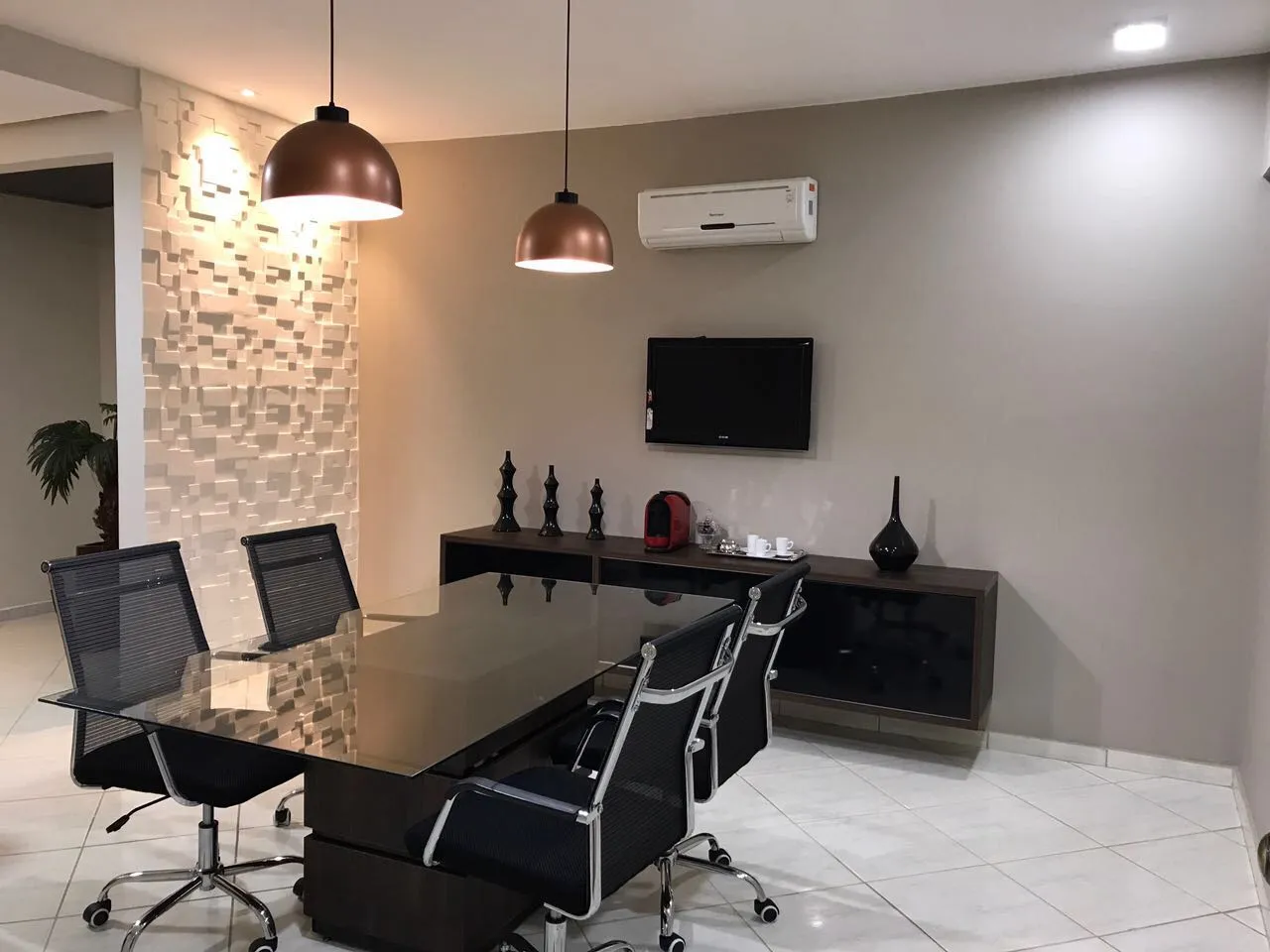 Sala de reuniões com mesa de vidro escuro, cadeiras pretas e luminárias pendentes